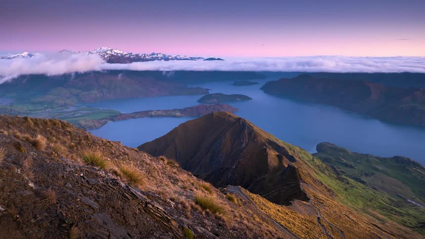 People walk on a mountain path as clouds move over the lake and there are snow covered mountains in the distance
