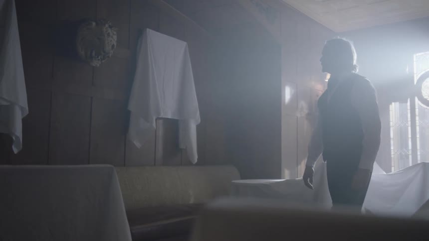 A man walks through a room with covered pictures and furniture and approaches a wall