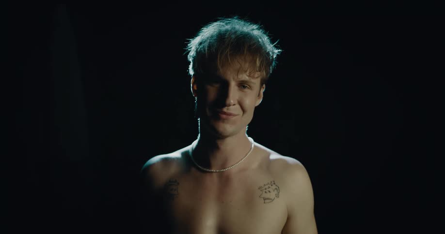 Topless man with tattoo and necklace kisses his biceps smiles at camera