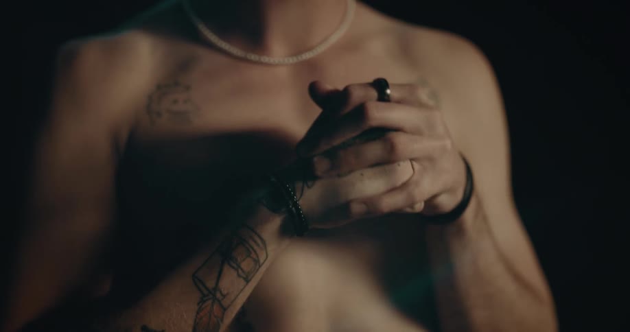 A man clasps his hands together while standing topless in a room