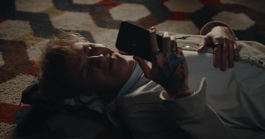 Man with white shirt and beige jacket lies on carpet using smartphone smiling