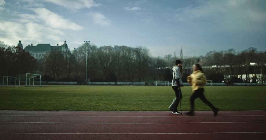 A woman walks along a track on crutches and stops as a runner passes by