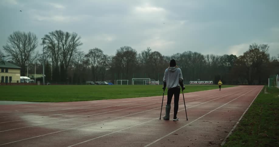 Woman with cast balances on crutches while watching jogger run on tracks