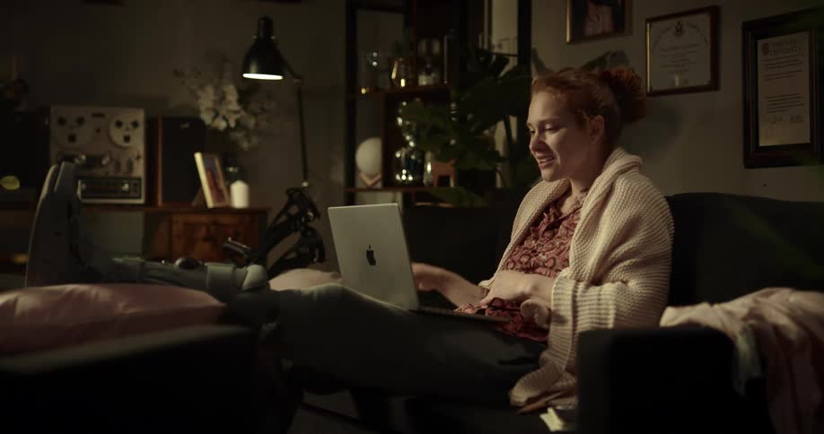 Young redheaded woman with boot splint sitting on couch leg elevated with laptop talking on zoom and examining papers