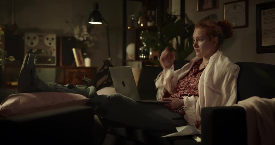 A woman lying on her bed with her laptop on her lap appearing to be confused about a conversation she is having she then picks up a piece of paper to help clarify her thoughts
