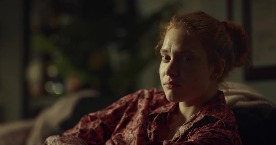Frowning redheaded woman struggles to smile slightly while sitting on couch