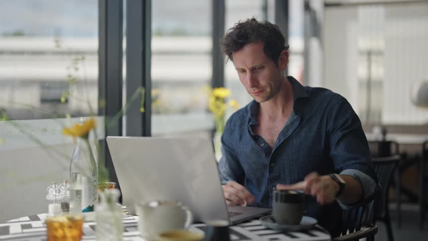 Man business casual sitting at restaurant table near window working on laptop and drinking coffee empty restaurant