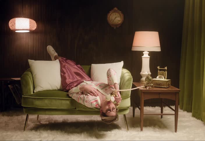 Blonde woman lies with her feet on back of loveseat and her legs hanging toward the rug as she talks on ornate rotary telephone