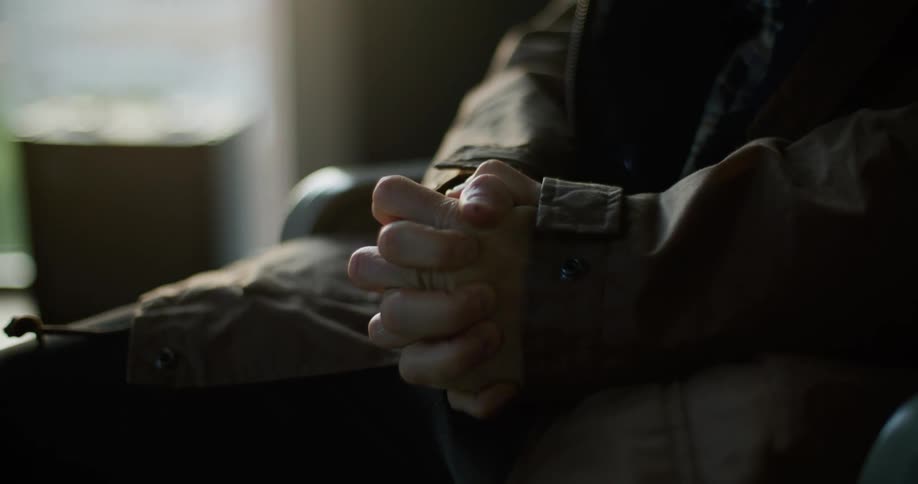 Man with jacket and pants sitting in armchair clasping hands together