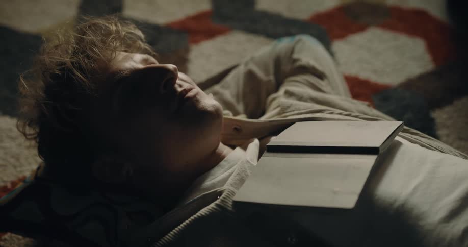 Man with white top lies on carpet with book on chest