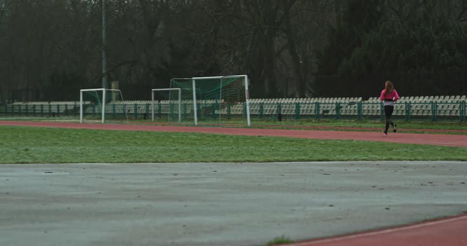A woman is running on a track field surrounded by trees under a cloudy sky