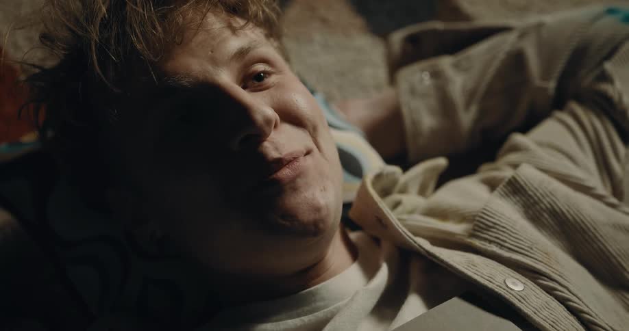 Man with brown jacket and white top lies on carpet looking up at ceiling and smiling in room