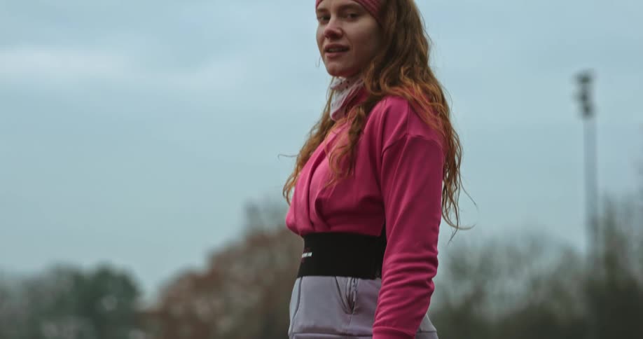 Woman stand on running track wearing gloves and a sweater smiles and waves