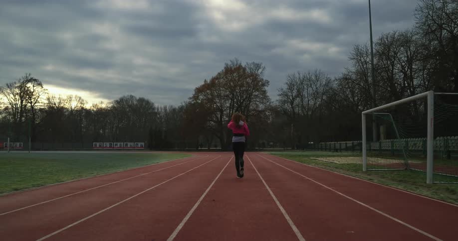A woman is running on a track field surrounded by trees under a cloudy sky