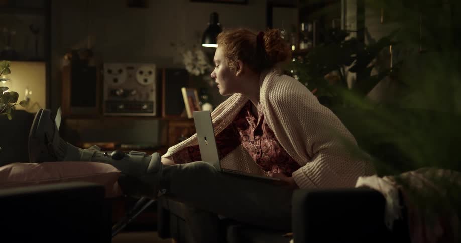 Young redheaded woman with leg cast sitting on couch with laptop picks up and examines an xray film probably of her leg or foot