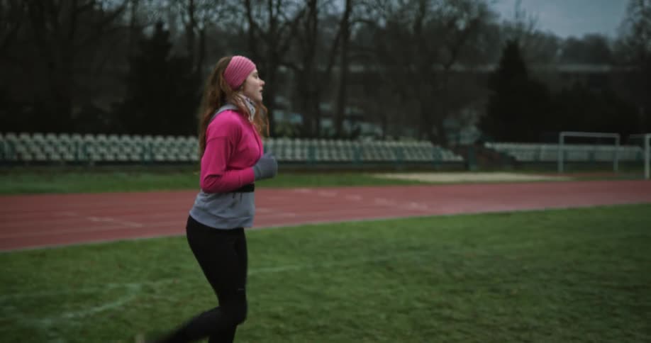 Young woman wearing pink workout gear warming up and running in place on the grass at a running track