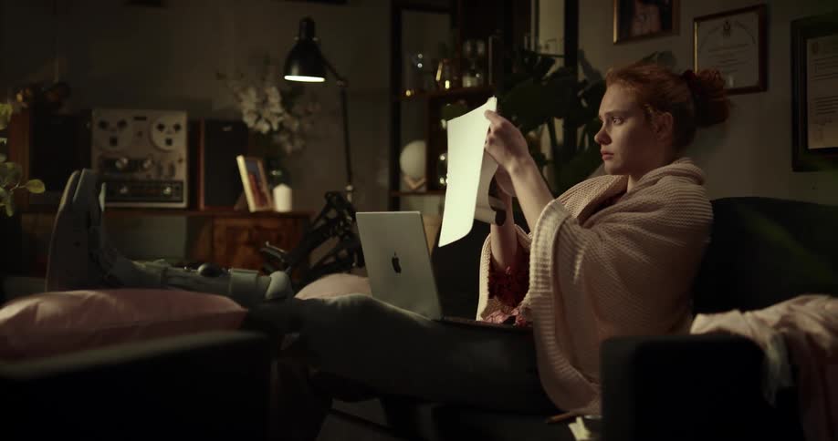 A lady on her bed with her laptop resting on her legs holding a book and opening it to read on a cozy night in