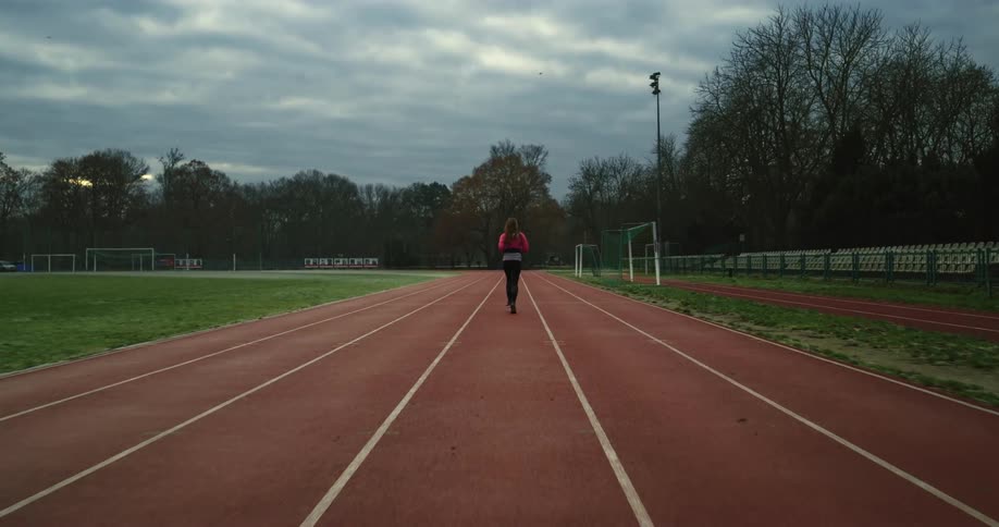 A woman is running on a track field surrounded by trees under a cloudy sky