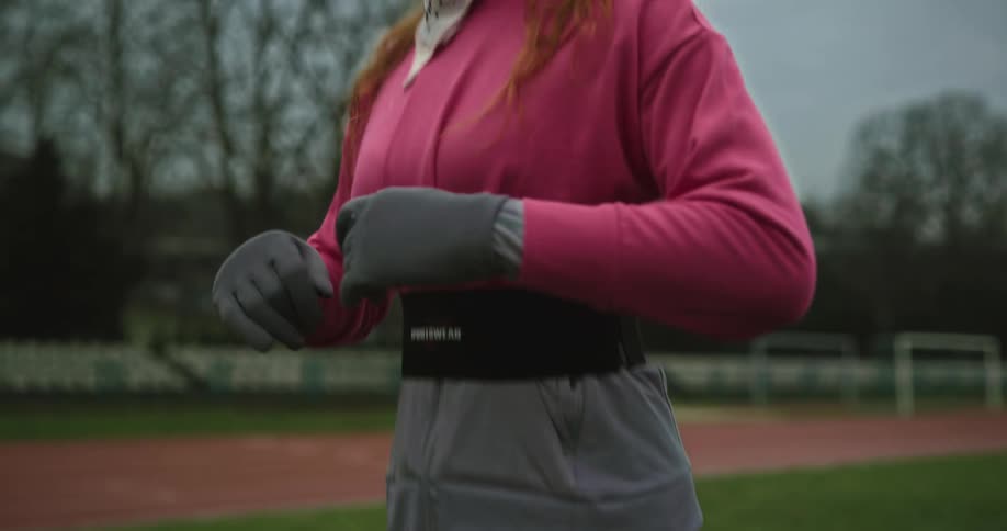 Woman with brown hair pink headband bandanna top black leggings and sneakers exercises on green lawn near running tracks