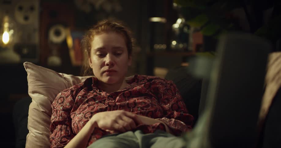 A teenage girl is sitting in a chair with a pillow behind her as she holds her phone and waves to someone with tears in her eyes and leans back with a sad face