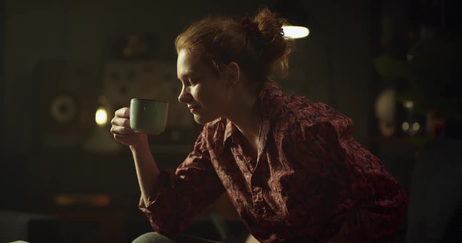 A woman sitting down is resting her head back on the chair as she sits up and drinks a cup of coffee