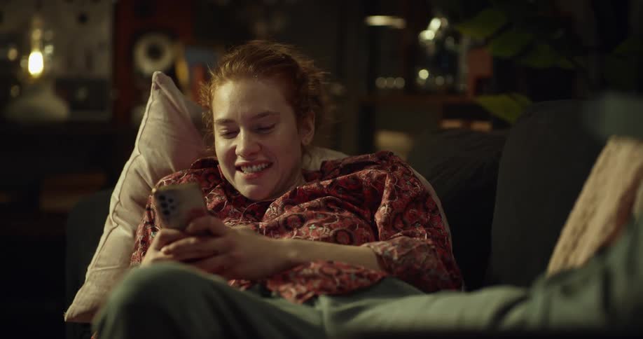 A woman smiles as she uses her smartphone while resting on a sofa