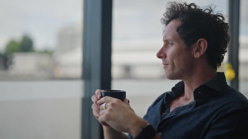 A man is sitting in a restaurant next to the windows drinking a cup of coffee