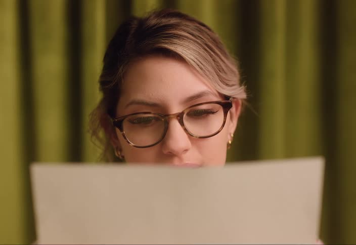 Woman with brown hair glasses earrings and white and red sweater reads a sheet of paper in room