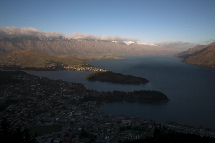 Queenstown, NZ is shown with several houses close together cars and buildings with a huge lake next to it and mountains in the background