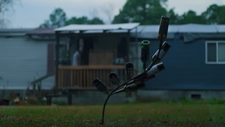 Man carries case up steps of mobile home with lawn ornament made from wine bottles and metal