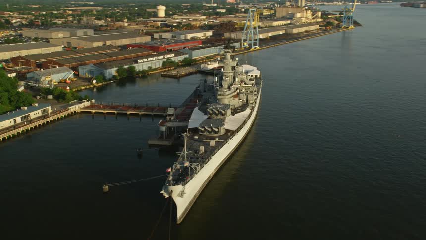 Aerial footage of the USS New Jersey docked at Camden New Jersey during sunset with industrial and military tags