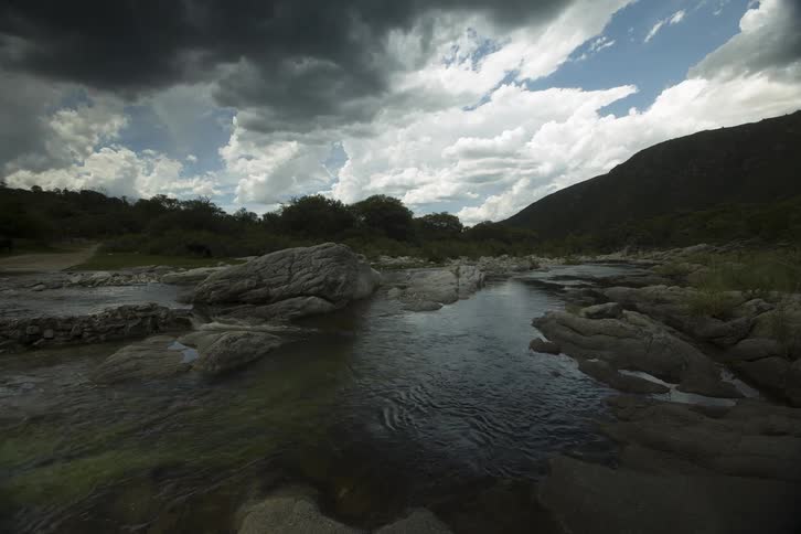 Timelapse of a clear running stream through rocks, as clouds float across the sky and there is a mountain and trees in the distance