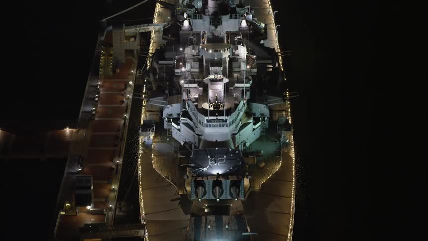 Aerial footage of the USS New Jersey in Camden NJ at night with the lights of the naval vessel illuminating the dark waters below
