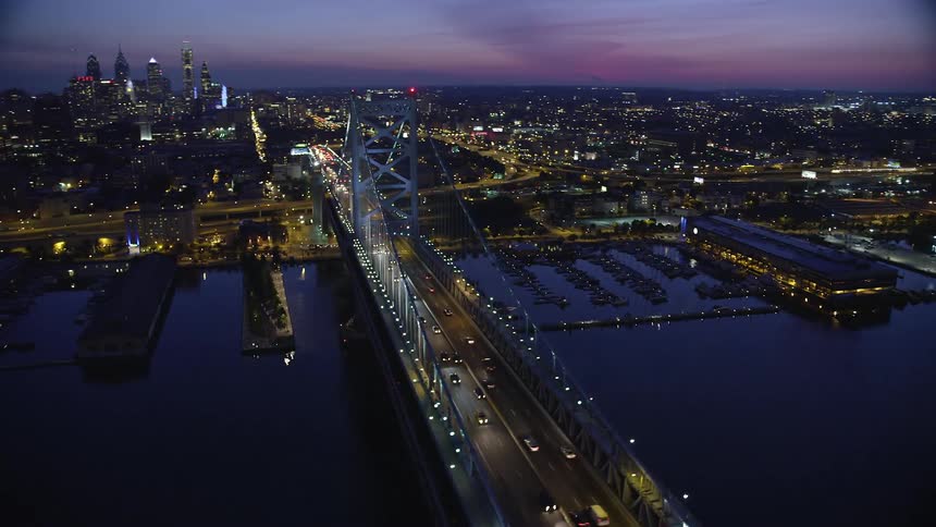 A stunning aerial view of Philadelphia at night reveals its impressive skyline and the Benjamin Franklin suspension bridge with traffic passing by while the partly cloudy sky adds drama to the clip