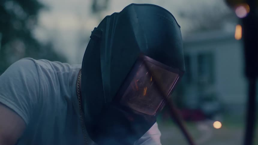 Man using welder while wearing welding face covering stops and lifts mask with house in background
