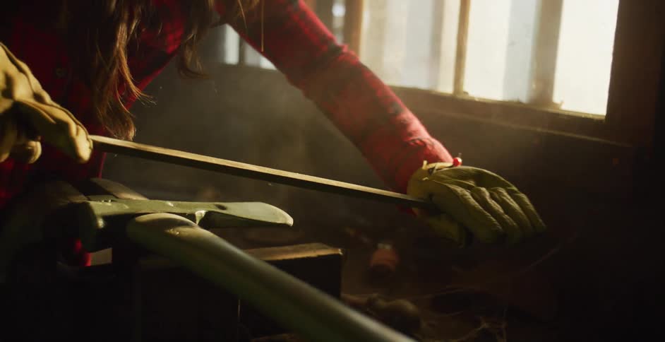 Woman with red plaid shirt and brown hair and gloves sharpens an ax