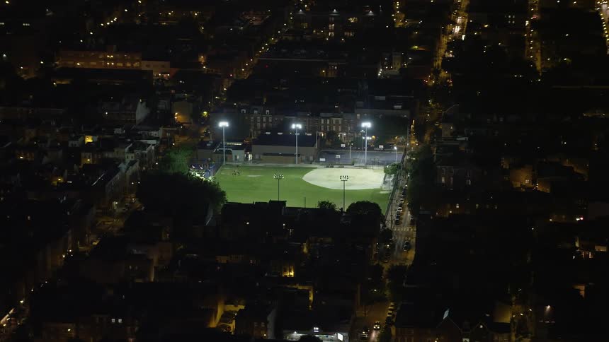Traffic flows along city streets at night past brightly lit empty baseball field