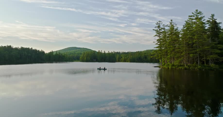 Two people are rowing a canoe across a river surrounded by forests and a mountain under a cloudy sky