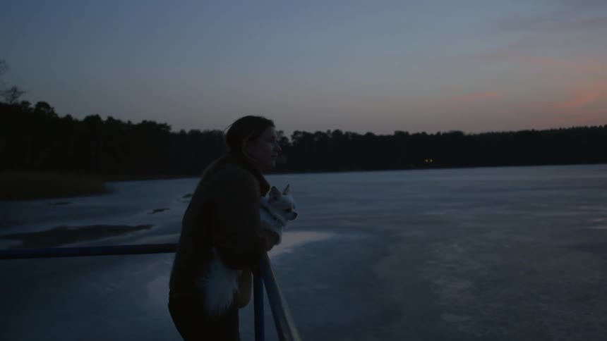 Woman holds dog while leaning against rail and looking out toward sea with forest covered shore in the background
