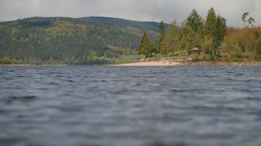 The water ripples and moves as a house along the river is surrounded by trees and there are mountains in the distance