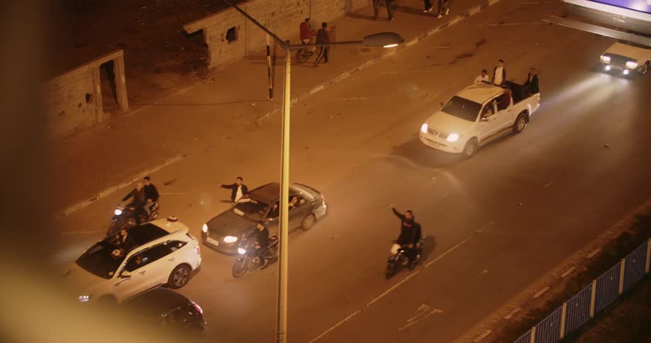 Cars vans and motorbikes drive down a street as pedestrians walk on the roadside