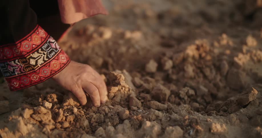 A woman with decorative patterns on her sleeve is picking up clumps of dirt and smashing them on the ground