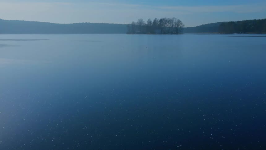 An aerial view flying over a large lake with a small island and surrounding mountains during the daytime