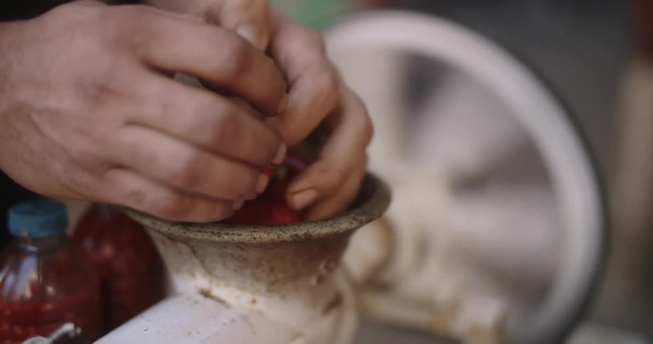 Closeup of mans hand putting meat into a meat grinder with the wheel in the background and two bottles of salsa