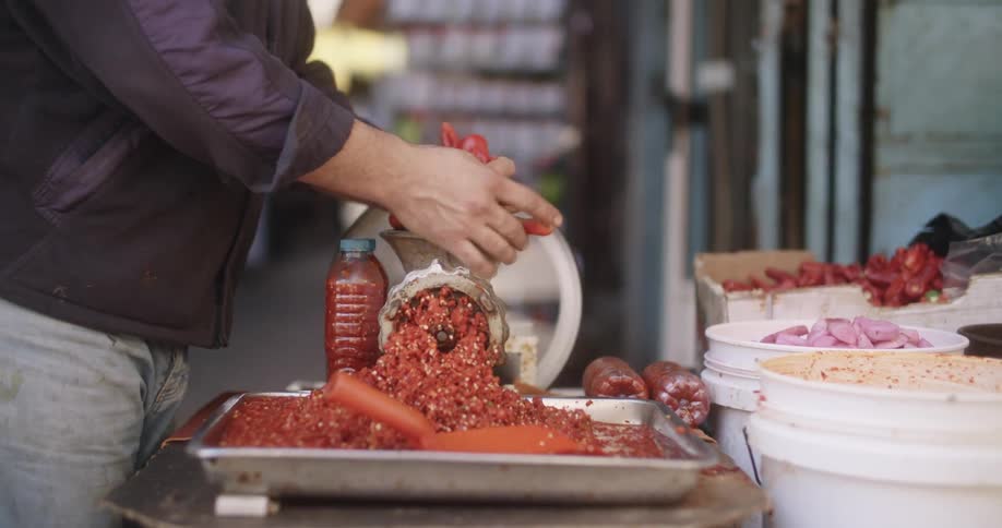 A human wearing a sweater grabbing from buckets of fresh produce specifically red hot peppers and putting into meat grinder presumably to make salsa