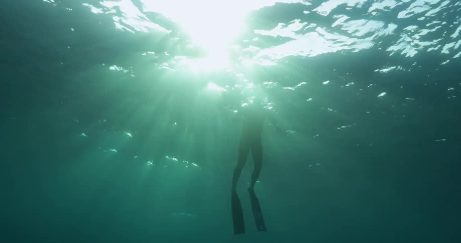 A scuba diver is swimming under water as sunlight shines brightly on the surface
