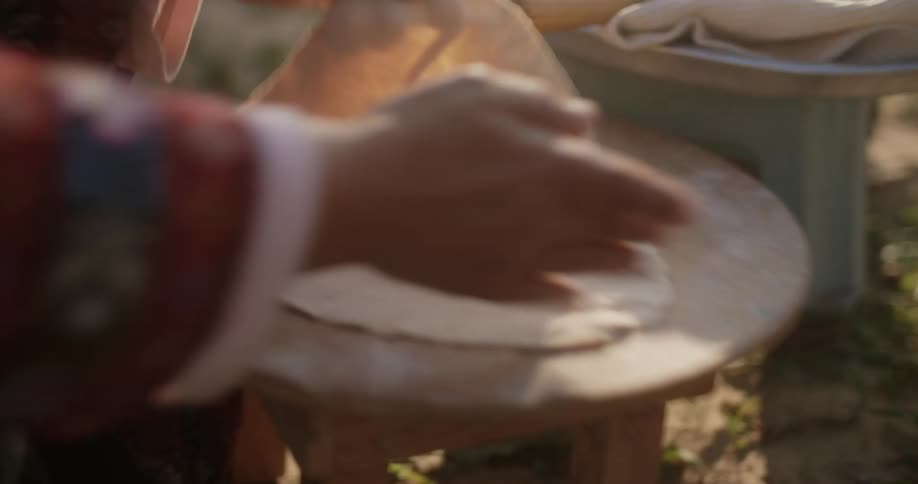 Woman cooks flatbread on heated dome stove before shaping another piece while outside