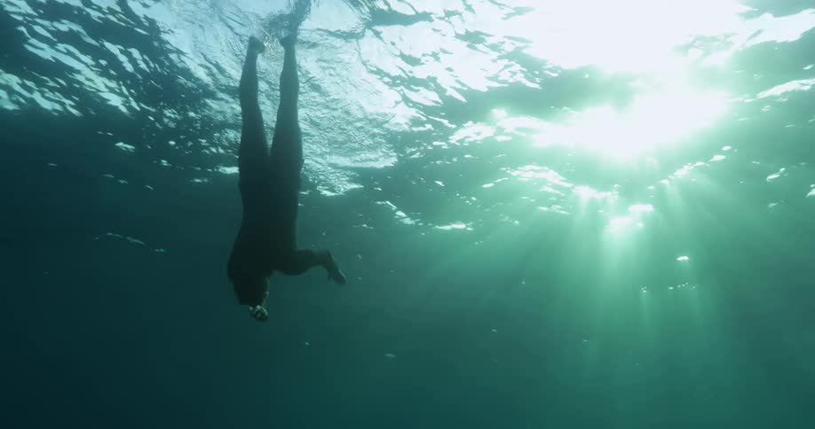 A scuba diver floating near the surface swims down deeper as sunlight shines brightly from above