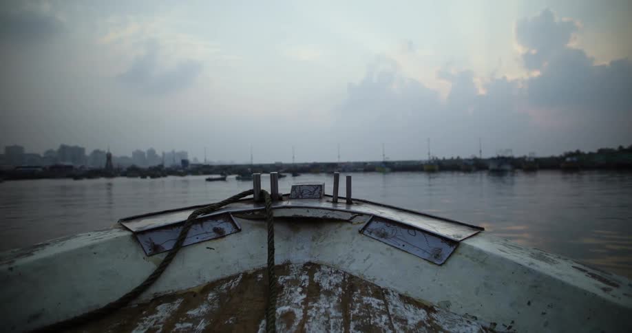 The front of an old metal boat with rust moves through the water towards the shore line to dock amoung the many other boats that are already docked as the sunsets below the clouded sky