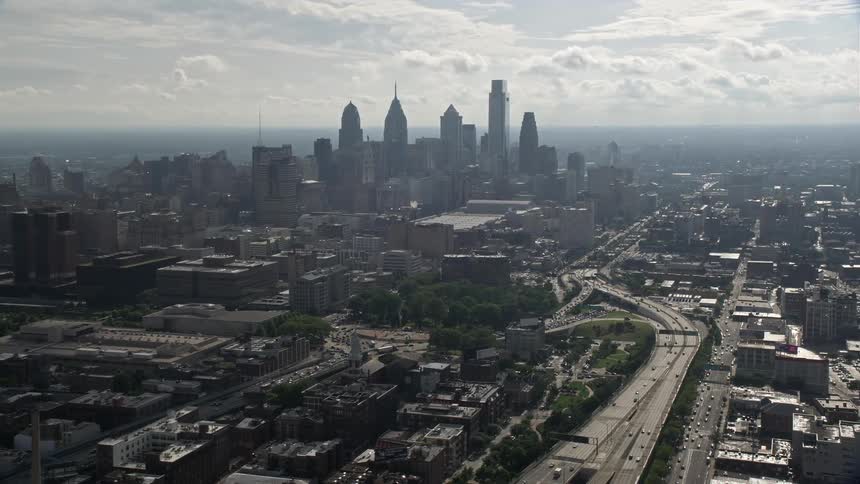 Aerial footage showing the Benjamin Franklin Suspension Bridge leading past a marina and over a highway towards the hazy downtown skyline of Philadelphia on a spring or summer evening